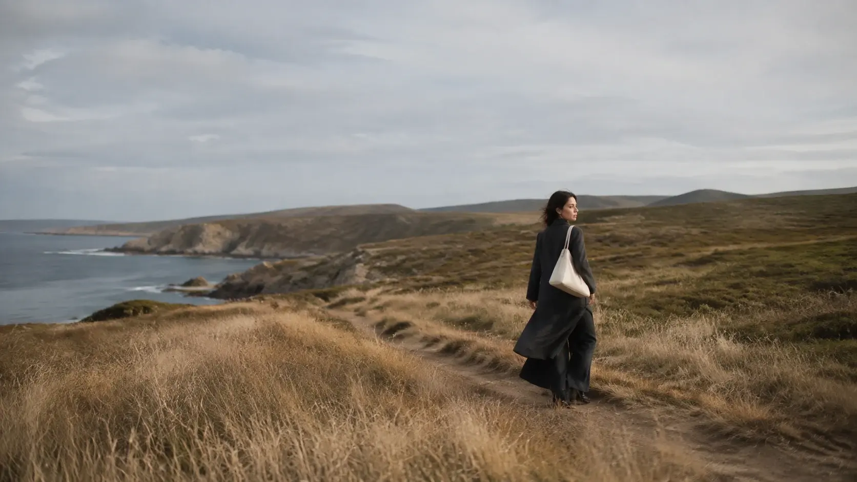 A woman with an ivory shoulder bag walks along a coastal path